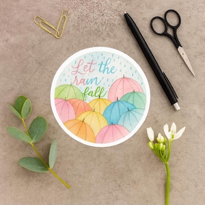 May include: A round sticker with the text "Let the rain fall" and watercolor umbrellas in various colors. The sticker is surrounded by a pen, scissors, a sprig of eucalyptus, a flower, and a paper clip.