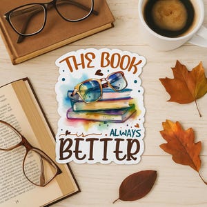 May include: A sticker with the text "THE BOOK is ALWAYS BETTER" in brown lettering. The sticker features a stack of books with glasses on top. A cup of coffee, glasses, and fall leaves are also in the image.