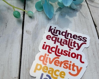 May include: A white sticker with the words "kindness, equality, inclusion, diversity, peace, love" in a rainbow of colors. The sticker is on a light wood surface with blue flowers.