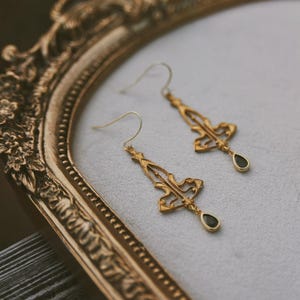 K&ouml;nnte beinhalten: Ein Paar goldene Ohrh&auml;nger mit schwarzen tropfenf&ouml;rmigen Steinen. Die Ohrringe haben ein zartes, filigranes Design.