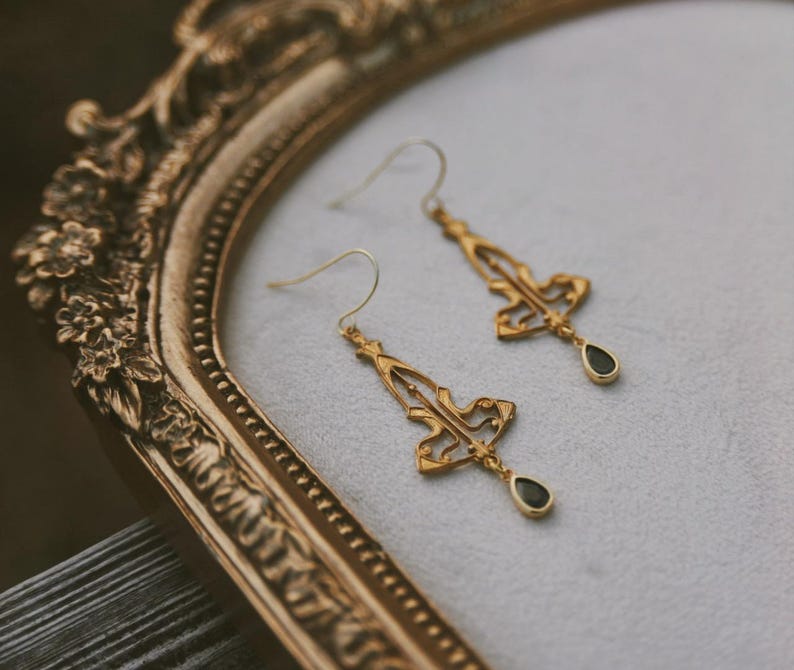 K&ouml;nnte beinhalten: Ein Paar goldene Ohrh&auml;nger mit schwarzen tropfenf&ouml;rmigen Steinen. Die Ohrringe haben ein zartes, filigranes Design.