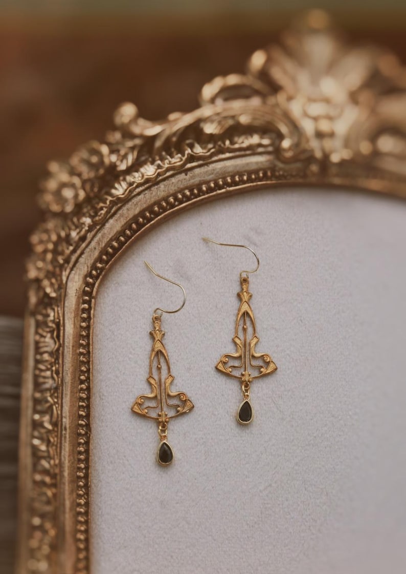 K&ouml;nnte beinhalten: Ein Paar goldene Ohrringe mit filigranen Designs und schwarzen Edelstein-Akzenten. Die Ohrringe werden auf einem goldfarbenen St&auml;nder pr&auml;sentiert.