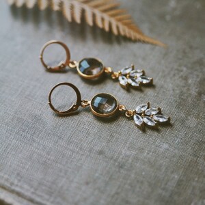 May include: A pair of gold earrings with a teardrop-shaped clear crystal and a round, smoky quartz stone. The earrings have a delicate, feminine design.
