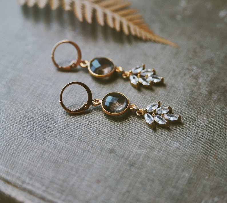 May include: A pair of gold earrings with a teardrop-shaped clear crystal and a round, smoky quartz stone. The earrings have a delicate, feminine design.