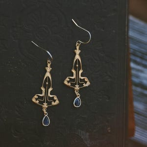 K&ouml;nnte beinhalten: Ein Paar goldene Ohrringe mit schwarzem Emaille-Design und einem tropfenf&ouml;rmigen blauen Edelstein-Anh&auml;nger. Die Ohrringe haben einen geometrischen Art-Deco-Stil.