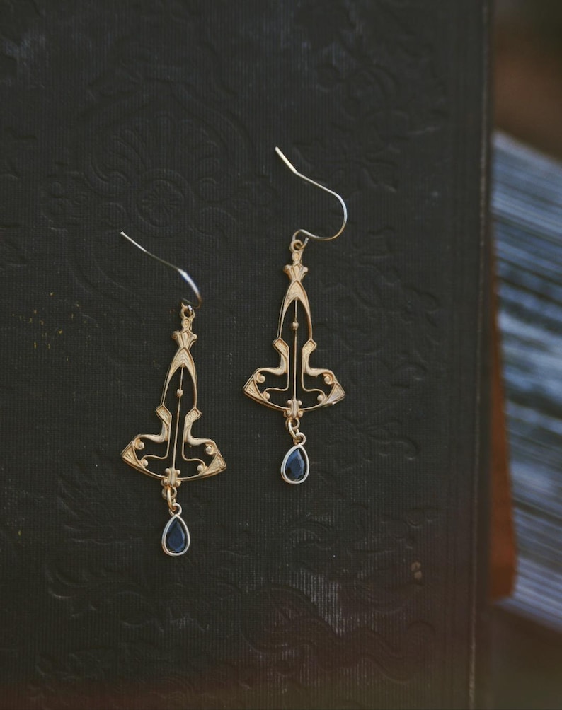 K&ouml;nnte beinhalten: Ein Paar goldene Ohrringe mit schwarzem Emaille-Design und einem tropfenf&ouml;rmigen blauen Edelstein-Anh&auml;nger. Die Ohrringe haben einen geometrischen Art-Deco-Stil.