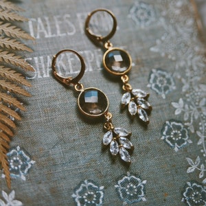 May include: A pair of gold earrings with a dark gray gemstone and clear crystal accents in the shape of leaves.