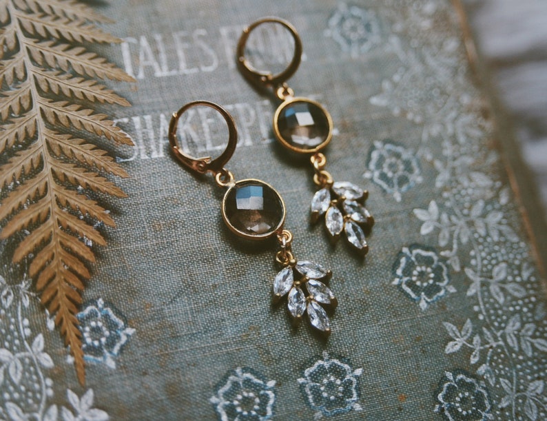 May include: A pair of gold earrings with a dark gray gemstone and clear crystal accents in the shape of leaves.