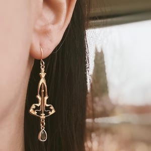 K&ouml;nnte beinhalten: Goldene Ohrh&auml;nger mit einem schwarzen tropfenf&ouml;rmigen Stein. Die Ohrringe haben ein filigranes Design mit einem geometrischen Muster.