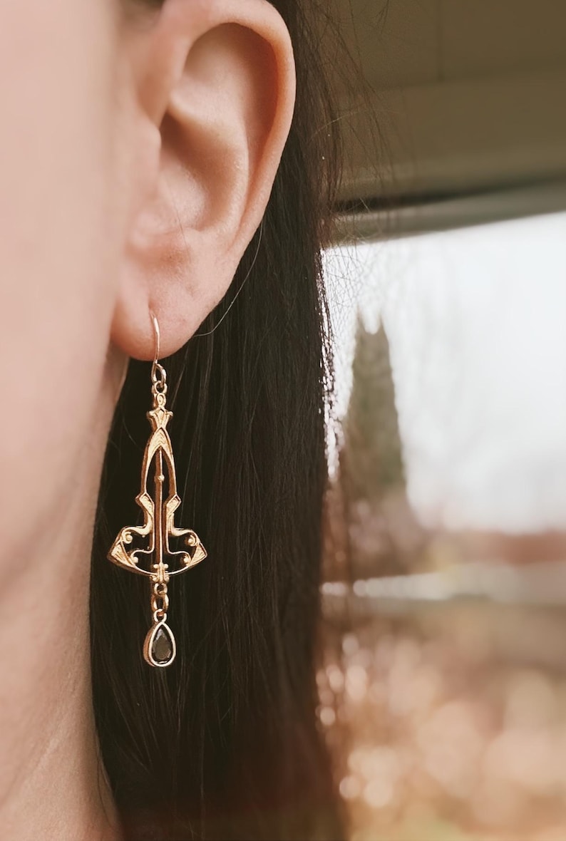 K&ouml;nnte beinhalten: Goldene Ohrh&auml;nger mit einem schwarzen tropfenf&ouml;rmigen Stein. Die Ohrringe haben ein filigranes Design mit einem geometrischen Muster.