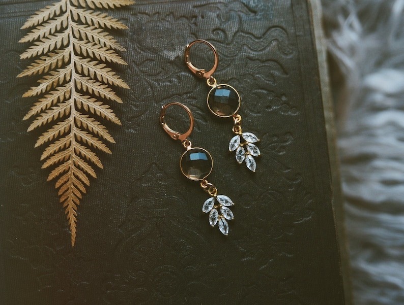 May include: A pair of gold earrings with a dark brown gemstone and clear crystal accents. The earrings are hanging from a dark surface.