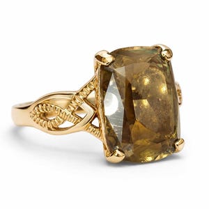 May include: A gold ring featuring a large, rectangular, olive-toned gemstone. The ring has an ornate, openwork design on the band and around the gemstone setting. The gemstone is faceted, catching the light.