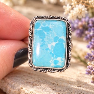 May include: A rectangular ring featuring a large, smooth, light blue stone with darker blue and white mottled patterns. The stone is set in a silver-toned metal frame with a twisted rope-like border design around the outer edge of the setting.