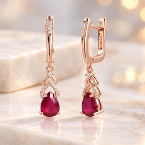 May include: A pair of rose gold drop earrings featuring pear-shaped red rubies suspended from a decorative frame accented with small clear stones. The hinged lever-back closures are stamped with S925, indicating sterling silver construction.