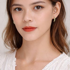 Pu&ograve; includere: Una donna indossa orecchini a cerchio dorati con piccole pietre preziose verdi. Gli orecchini sono incastonati in un metallo dorato e hanno la forma di un semicerchio. La donna indossa un top di pizzo bianco.