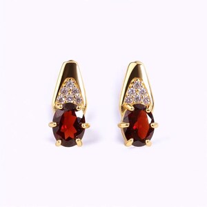 May include: A pair of gold-colored earrings. Each earring features a large, oval-shaped, deep red gemstone, accented with a triangle of small, clear stones and gold-colored metal. The earrings have a post-back closure.