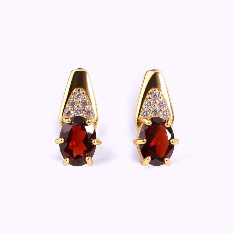 May include: A pair of gold-colored earrings. Each earring features a large, oval-shaped, deep red gemstone, accented with a triangle of small, clear stones and gold-colored metal. The earrings have a post-back closure.