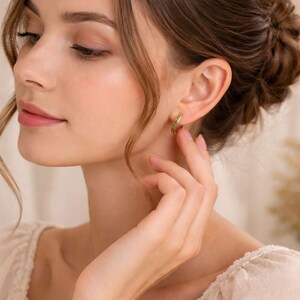 Pu&ograve; includere: Una donna indossa orecchini a cerchio dorati con piccole pietre verdi. Gli orecchini hanno un design semplice ed elegante. La donna indossa una camicetta color crema con dettagli in pizzo.