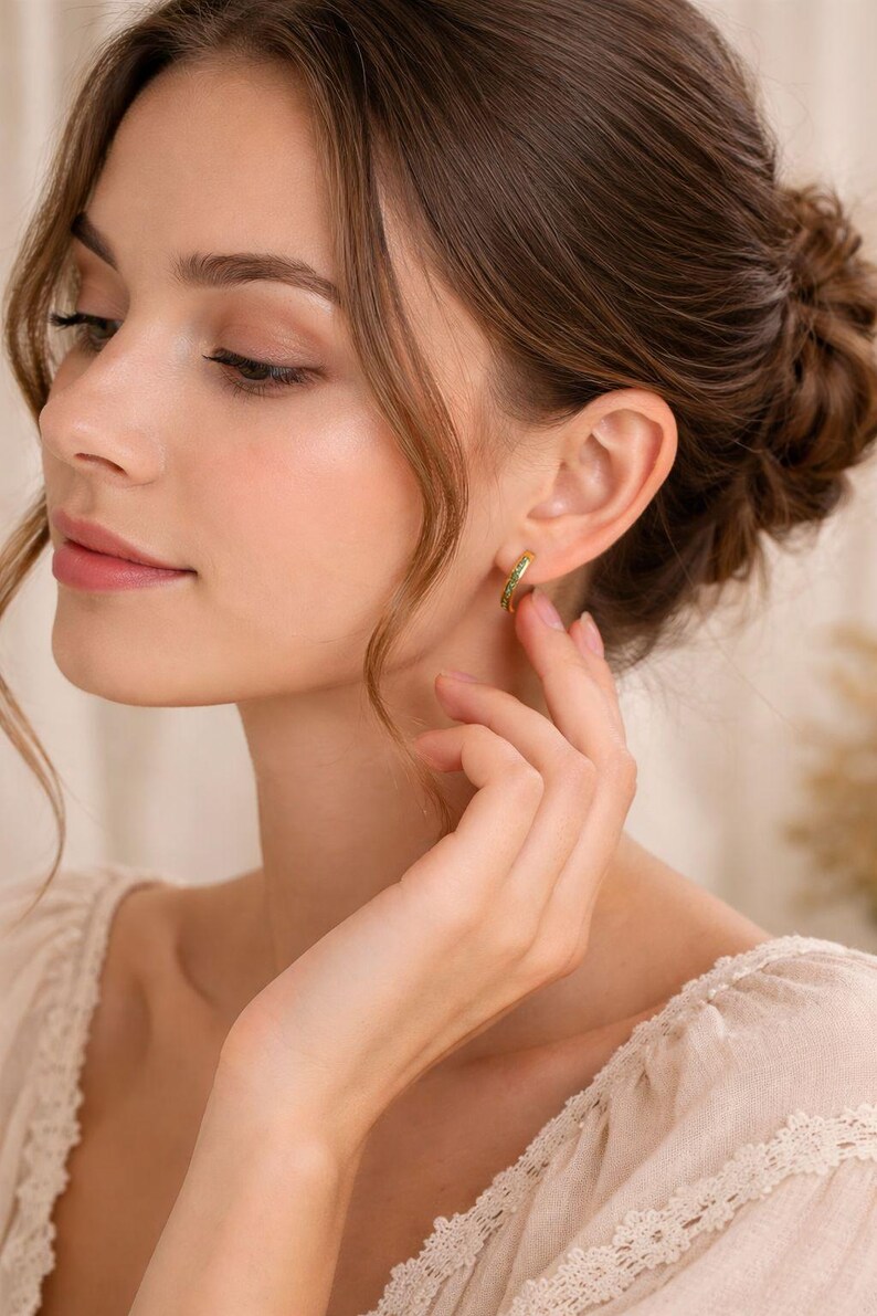 Pu&ograve; includere: Una donna indossa orecchini a cerchio dorati con piccole pietre verdi. Gli orecchini hanno un design semplice ed elegante. La donna indossa una camicetta color crema con dettagli in pizzo.