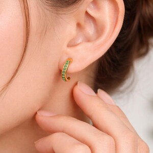 Pu&ograve; includere: Orecchini a cerchio color oro con una fila di piccole pietre preziose verdi. Gli orecchini sono indossati su un orecchio, con una mano che tocca delicatamente la guancia. Design curvo.