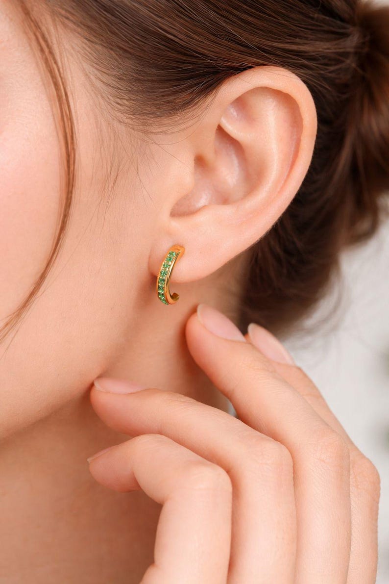 Pu&ograve; includere: Orecchini a cerchio color oro con una fila di piccole pietre preziose verdi. Gli orecchini sono indossati su un orecchio, con una mano che tocca delicatamente la guancia. Design curvo.