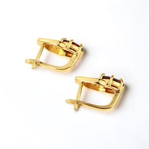 May include: A pair of gold-colored earrings with a lever back closure. Each earring features three rectangular, deep red gemstones set in a gold frame. The earrings are displayed on a white background.