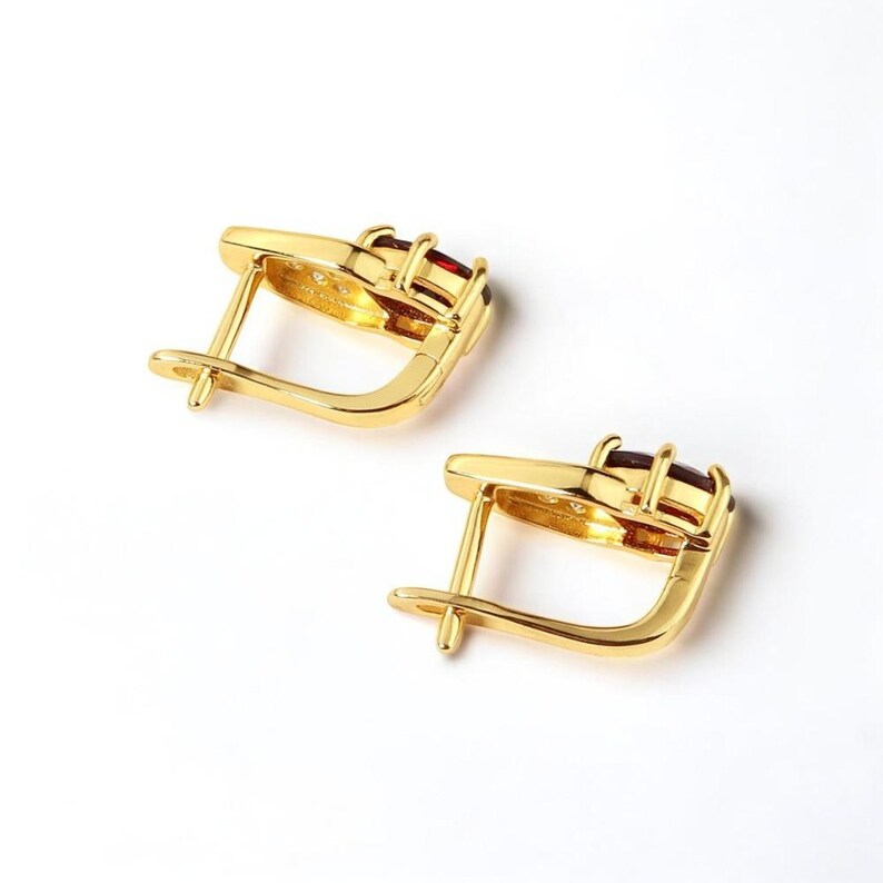 May include: A pair of gold-colored earrings with a lever back closure. Each earring features three rectangular, deep red gemstones set in a gold frame. The earrings are displayed on a white background.