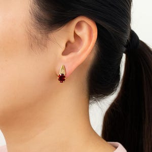 May include: A close-up of a gold earring with a teardrop design. The earring features a large, oval-shaped, deep red gemstone at the bottom and a small, clear stone at the top. The earring is worn on a person's ear.
