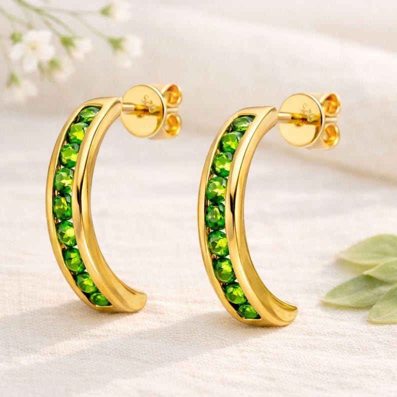 Pu&ograve; includere: Un paio di orecchini a cerchio color oro. Ogni orecchino ha una forma curva, foderata con piccole pietre preziose rotonde di colore verde brillante. Gli orecchini hanno un perno e un retro per un uso sicuro. Lo sfondo &egrave; in morbido tessuto bianco.