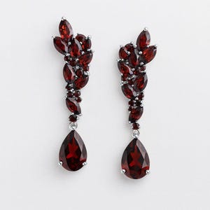 Puede incluir: Un par de elegantes pendientes colgantes con racimos de piedras de color rojo oscuro con corte marquesa y una gran piedra en forma de lágrima en la parte inferior. Los pendientes tienen un engaste de metal plateado.