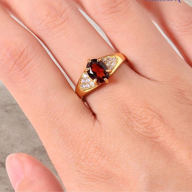 May include: A gold ring featuring a large, oval-cut, deep red gemstone as the centerpiece. The ring is flanked by smaller, clear gemstones on each side, set in a gold band. The ring is a piece of jewelry.