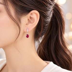 May include: A silver drop earring featuring a vibrant oval-cut red gemstone suspended from a slender, vertical bar set with small, clear, sparkling stones. The earring is secured with a hinged clasp mechanism, showcasing a polished metallic finish.