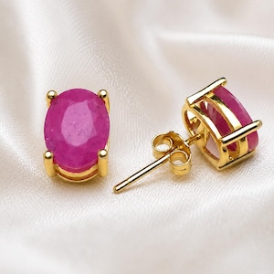 Op de afbeelding: Een paar gouden oorstekers met ovale, levendige roze edelstenen. De edelstenen zijn gezet in een gouden bezel setting, gepresenteerd op een witte, zijdeachtige ondergrond. Klassieke sieraden.