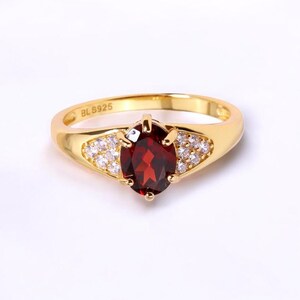 May include: A gold-toned ring featuring a large, oval-cut, deep red gemstone as the centerpiece. The ring is further embellished with smaller, clear gemstones on either side of the central stone. The band is smooth and polished, with the inscription "BL S925".
