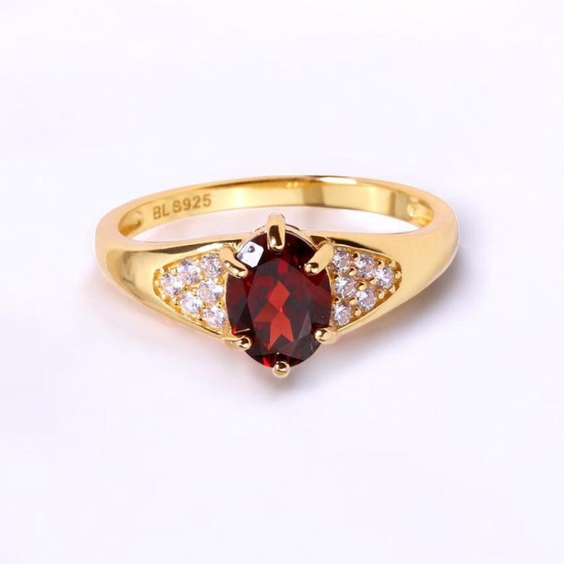 May include: A gold-toned ring featuring a large, oval-cut, deep red gemstone as the centerpiece. The ring is further embellished with smaller, clear gemstones on either side of the central stone. The band is smooth and polished, with the inscription "BL S925".