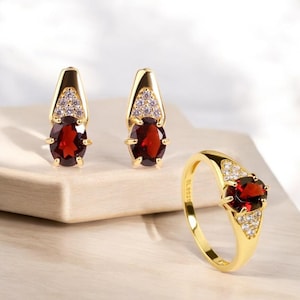 May include: A gold-toned jewelry set featuring a ring and a pair of earrings. Each piece showcases a large, oval, deep red gemstone, complemented by smaller, clear stones. The ring and earrings have a classic, elegant design.