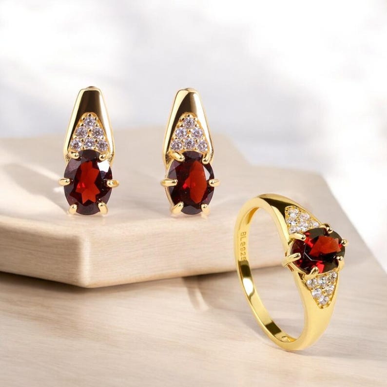 May include: A gold-toned jewelry set featuring a ring and a pair of earrings. Each piece showcases a large, oval, deep red gemstone, complemented by smaller, clear stones. The ring and earrings have a classic, elegant design.