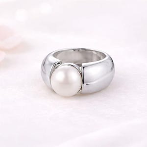 Puede incluir: Un anillo de metal plateado pulido con una perla blanca redonda y lisa engastada en el centro de una banda ancha y curva. La superficie metálica es reflectante y lisa, creando un aspecto limpio y continuo que resalta el brillo de la perla.