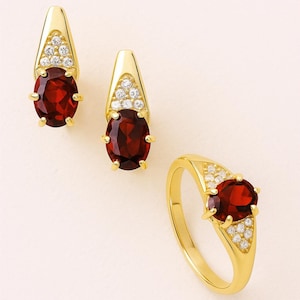May include: A gold-toned jewelry set featuring a ring and a pair of earrings. Each piece has a large, oval, deep red gemstone and is accented with small, clear gemstones. The earrings have a triangular top.