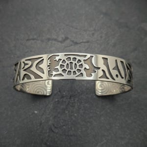 May include: Silver bracelet with a cutout design of a turtle and other abstract shapes. The bracelet is open and has a smooth, polished finish.