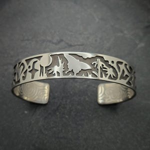 May include: A silver bracelet with a cutout design featuring various shapes and symbols. The bracelet is open and has a smooth, polished finish.