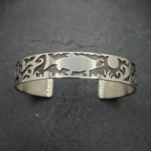May include: A silver bracelet with a cutout design of a fish and other aquatic imagery. The bracelet is made of sterling silver and has a polished finish.
