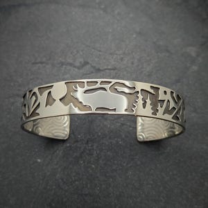 May include: A silver bracelet with a cut-out design of a deer and trees. The bracelet is on a gray background.