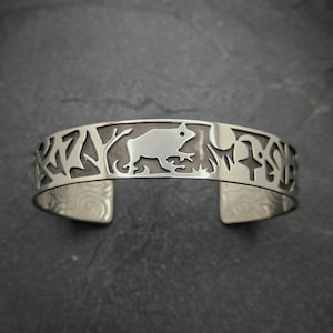 May include: Silver cuff bracelet featuring a cut-out design of a frog, trees, and other natural elements. The open-ended bracelet has a polished finish and is designed to be worn on the wrist. The bracelet is approximately 2.5 inches wide.