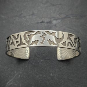 May include: A silver bracelet with a cutout design of stylized figures. The bracelet has a textured finish.