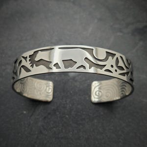 May include: A silver bracelet with a cutout design of a stylized animal and floral elements. The bracelet has a textured finish on the inside.