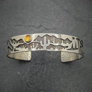 May include: A silver bracelet with a cut-out design of a desert landscape, including mountains, cacti, and a sun. A small yellow gemstone is set in the center of the bracelet, representing the sun.