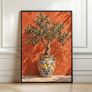 Op de afbeelding: Een ingelijste kunstprint met een olijfboom met groene en donkere olijven in een decoratieve keramische pot. De pot is beschilderd met gele citroenen, blauwe bladeren en oranje bloemen. De achtergrond is een getextureerde, levendige oranje muur.