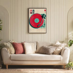 Op de afbeelding: Een ingelijste poster met een rode vinylplaat op een platenspeler, met Japanse tekst linksboven. De poster heeft een cr&egrave;mekleurige achtergrond en wordt weergegeven boven een beige bank met decoratieve kussens.