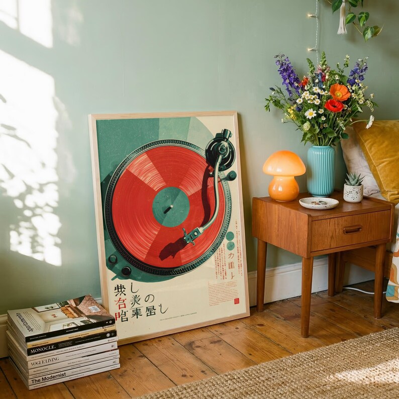 May include: A framed poster featuring a graphic illustration of a turntable with a vibrant red vinyl record. The artwork includes Japanese text and a stylized green background. The poster is positioned on a wooden floor next to a stack of magazines.